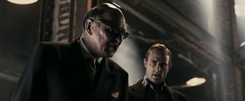 Movie still from “RocknRolla” (2008), directed by Guy Ritchie – A man in a suit and tie standing next to another man in a suit and tie; Medium shot, Over the shoulder angle