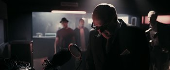 Movie still from “RocknRolla” (2008), directed by Guy Ritchie – A man in a suit and glasses looking at a microphone; Medium shot, Low angle