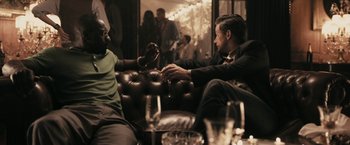 Movie still from “RocknRolla” (2008), directed by Guy Ritchie – Two men sitting on a couch holding wine glasses; Medium shot, Over the shoulder angle