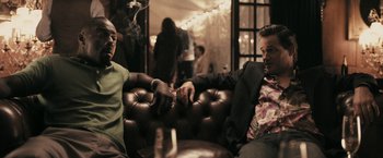 Movie still from “RocknRolla” (2008), directed by Guy Ritchie – Two men sitting on a leather couch and one is smoking a cigarette; Medium shot, Low angle