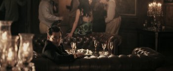 Movie still from “RocknRolla” (2008), directed by Guy Ritchie – A man sitting on a couch in front of a group of people; Medium shot, Over the shoulder angle
