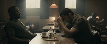 Movie still from “RocknRolla” (2008), directed by Guy Ritchie – A man sitting at a table with his head down in front of a cup; Medium shot, Low angle