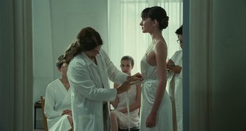 Movie still from “Saint Laurent” (2014), directed by Bertrand Bonello – A woman in a white lab coat is measuring a woman's waist; Medium shot, Over the shoulder angle