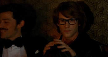 Movie still from “Saint Laurent” (2014), directed by Bertrand Bonello – A young man wearing glasses is sitting in a room; Close Up shot, Over the shoulder angle