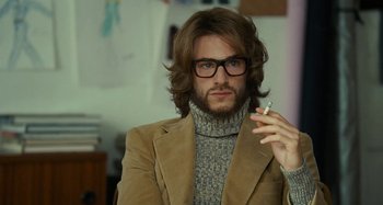 Movie still from “Saint Laurent” (2014), directed by Bertrand Bonello – A man in a brown jacket smoking a cigarette in a room; Close Up shot, Over the shoulder angle