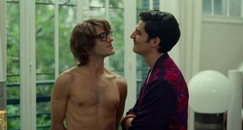 Movie still from “Saint Laurent” (2014), directed by Bertrand Bonello – Two young men standing next to each other in front of a window; Medium shot, Over the shoulder angle