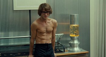Movie still from “Saint Laurent” (2014), directed by Bertrand Bonello – A shirtless man standing in front of a table; Medium shot, Over the shoulder angle