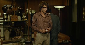 Movie still from “Saint Laurent” (2014), directed by Bertrand Bonello – A man in a brown shirt is standing in a living room; Medium shot, Over the shoulder angle
