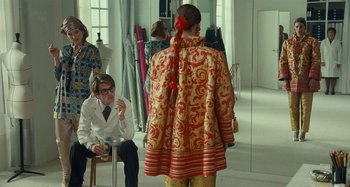Movie still from “Saint Laurent” (2014), directed by Bertrand Bonello – A man in a white lab coat is looking at a woman in a red and gold coat; Medium shot, Over the shoulder angle