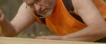 Movie still from “Sandpaper” (2018), directed by Jarett Bellucci – An older man in an orange shirt working on a table; Close Up shot, Over the shoulder angle