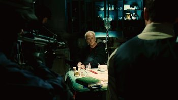 Movie still from “Saw II” (2005), directed by Darren Lynn Bousman – An older man sitting at a table in front of a camera; Medium shot, Over the shoulder angle