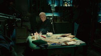 Movie still from “Saw II” (2005), directed by Darren Lynn Bousman – An older man sitting at a table writing on a piece of paper; Wide shot, High angle