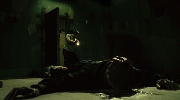Movie still from “Saw II” (2005), directed by Darren Lynn Bousman – A man standing next to a dead body in a dark room; Wide shot, High angle