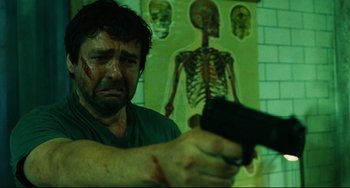 Movie still from “Saw III” (2006), directed by Darren Lynn Bousman – A man holding a gun in front of a wall with an image on it; Close Up shot, Low angle