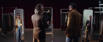 Movie still from “Official Competition” (2021), directed by Gastón Duprat – Two men are standing next to each other in front of a row of clothing racks; Medium shot, Over the shoulder angle