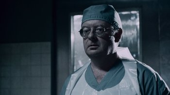 Movie still from “Saw IV” (2007), directed by Darren Lynn Bousman – A man wearing a surgical gown and glasses in a room; Close Up shot, Low angle