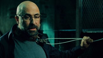 Movie still from “Saw V” (2008), directed by David Hackl – A bald man with glasses and a beard is holding a pair of headphones; Close Up shot, Over the shoulder angle
