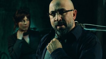 Movie still from “Saw V” (2008), directed by David Hackl – A man with a goatee and glasses is adjusting his neck tie; Close Up shot, Over the shoulder angle