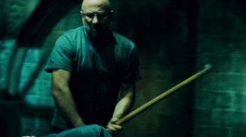 Movie still from “Saw V” (2008), directed by David Hackl – A man holding a wooden stick while sitting on a chair; Medium shot, Over the shoulder angle
