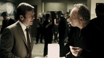 Movie still from “Saw VI” (2009), directed by Kevin Greutert – Two men in suits talking to each other in front of a group of people; Medium shot, Over the shoulder angle