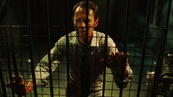 Movie still from “Saw VI” (2009), directed by Kevin Greutert – A man standing behind a metal fence with his hands on his hips; Medium shot, Low angle