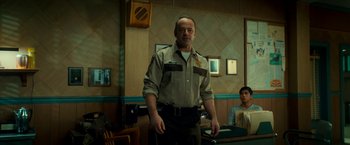 Movie still from “Scary Stories to Tell in the Dark” (2019), directed by André Øvredal – A man standing in front of a wall with two people sitting in the background; Medium shot, Over the shoulder angle