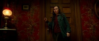 Movie still from “Scary Stories to Tell in the Dark” (2019), directed by André Øvredal – A woman standing in front of a door wearing glasses; Medium shot, Over the shoulder angle