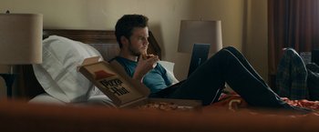 Movie still from “Scream” (2022), directed by Matt Bettinelli-Olpin – A man sitting on a bed eating a slice of pizza; Medium shot, High angle