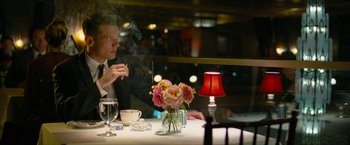 Movie still from “Seberg” (2019), directed by Benedict Andrews – A man sitting at a table with a vase filled with flowers; Medium shot, Over the shoulder angle