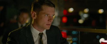 Movie still from “Seberg” (2019), directed by Benedict Andrews – A man wearing a suit and tie sitting in front of some lights; Close Up shot, Over the shoulder angle