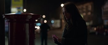 Movie still from “Official Secrets” (2019), directed by Gavin Hood – A woman standing in front of a red mailbox holding a pink card; Medium shot, Over the shoulder angle