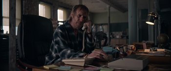 Movie still from “Official Secrets” (2019), directed by Gavin Hood – A man sitting at a table talking on a phone; Medium shot, Over the shoulder angle