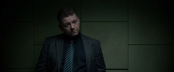 Movie still from “Official Secrets” (2019), directed by Gavin Hood – A man wearing a suit and tie in front of a wall; Medium shot, Over the shoulder angle