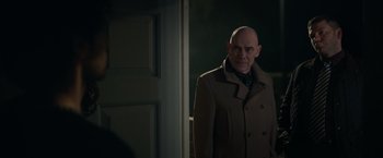 Movie still from “Official Secrets” (2019), directed by Gavin Hood – A bald man in a brown coat standing in front of a door; Medium shot, Over the shoulder angle