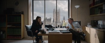 Movie still from “Official Secrets” (2019), directed by Gavin Hood – Two people sitting at a table in front of a large window; Medium shot, Over the shoulder angle