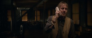 Movie still from “Official Secrets” (2019), directed by Gavin Hood – A man talking on a cell phone while wearing a brown vest; Medium shot, Over the shoulder angle