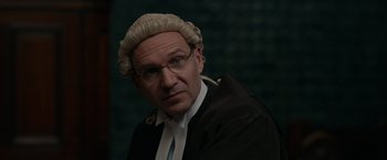 Movie still from “Official Secrets” (2019), directed by Gavin Hood – A man wearing a wig and glasses in a court room; Close Up shot, Over the shoulder angle