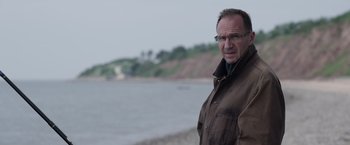 Movie still from “Official Secrets” (2019), directed by Gavin Hood – A man standing on a beach near a body of water; Close Up shot, Over the shoulder angle
