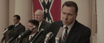 Movie still from “Selma” (2014), directed by Ava DuVernay – A man in a suit and tie standing in front of microphones in front of a confederate flag; Close Up shot, Over the shoulder angle
