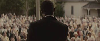 Movie still from “Selma” (2014), directed by Ava DuVernay – A man in a black suit is looking at a crowd of onlookers; Medium shot, Over the shoulder angle