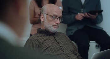 Movie still from “She Said” (2022), directed by Maria Schrader – An older man wearing glasses and a plaid shirt; Medium shot, Over the shoulder angle