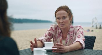 Movie still from “She Said” (2022), directed by Maria Schrader – A woman sitting at a table on the beach with two cups; Medium shot, Over the shoulder angle