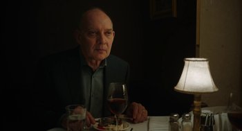 Movie still from “She Said” (2022), directed by Maria Schrader – An older man sitting at a dinner table with a glass of wine; Medium shot, Over the shoulder angle