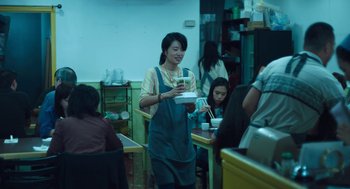 Movie still from “She Said” (2022), directed by Maria Schrader – A woman holding a tray of food in front of a group of people; Medium shot, Over the shoulder angle