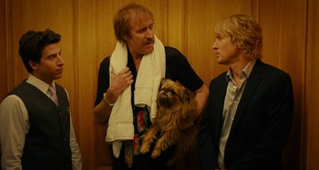 Movie still from “She's Funny That Way” (2014), directed by Peter Bogdanovich – A man holding a dog while standing next to another man; Medium shot, Low angle