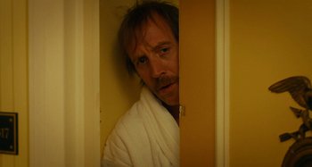Movie still from “She's Funny That Way” (2014), directed by Peter Bogdanovich – A man in a white bathrobe peeks out from behind a door; Close Up shot, Over the shoulder angle