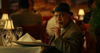 Movie still from “She's Funny That Way” (2014), directed by Peter Bogdanovich – An older man sitting at a table in front of papers; Close Up shot, Over the shoulder angle