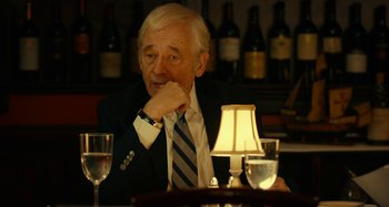Movie still from “She's Funny That Way” (2014), directed by Peter Bogdanovich – An older man sitting at a table in front of wine glasses; Close Up shot, Over the shoulder angle