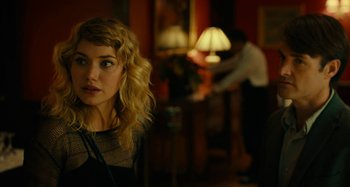 Movie still from “She's Funny That Way” (2014), directed by Peter Bogdanovich – A woman with blonde curly hair looking at the camera with a man in the background; Close Up shot, Over the shoulder angle
