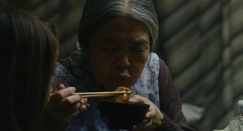 Movie still from “Shoplifters” (2018), directed by Kore-eda Hirokazu – An older woman eating food with chopsticks in a bowl; Close Up shot, Over the shoulder angle
